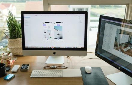 Masada duran, ekranında tasarım yazılımı açık olan bir iMac bilgisayar ve yanında başka bir monitörün de bulunduğu bir çalışma alanı. Masada ayrıca klavye, fare, akıllı saat, bazı küçük kutular, bir kedi heykeli ve küçük bir bitki bulunmaktadır.